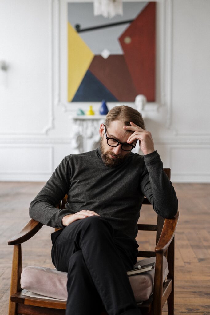 A man sitting in a chair with one hand on forehead looking down in deep thought.